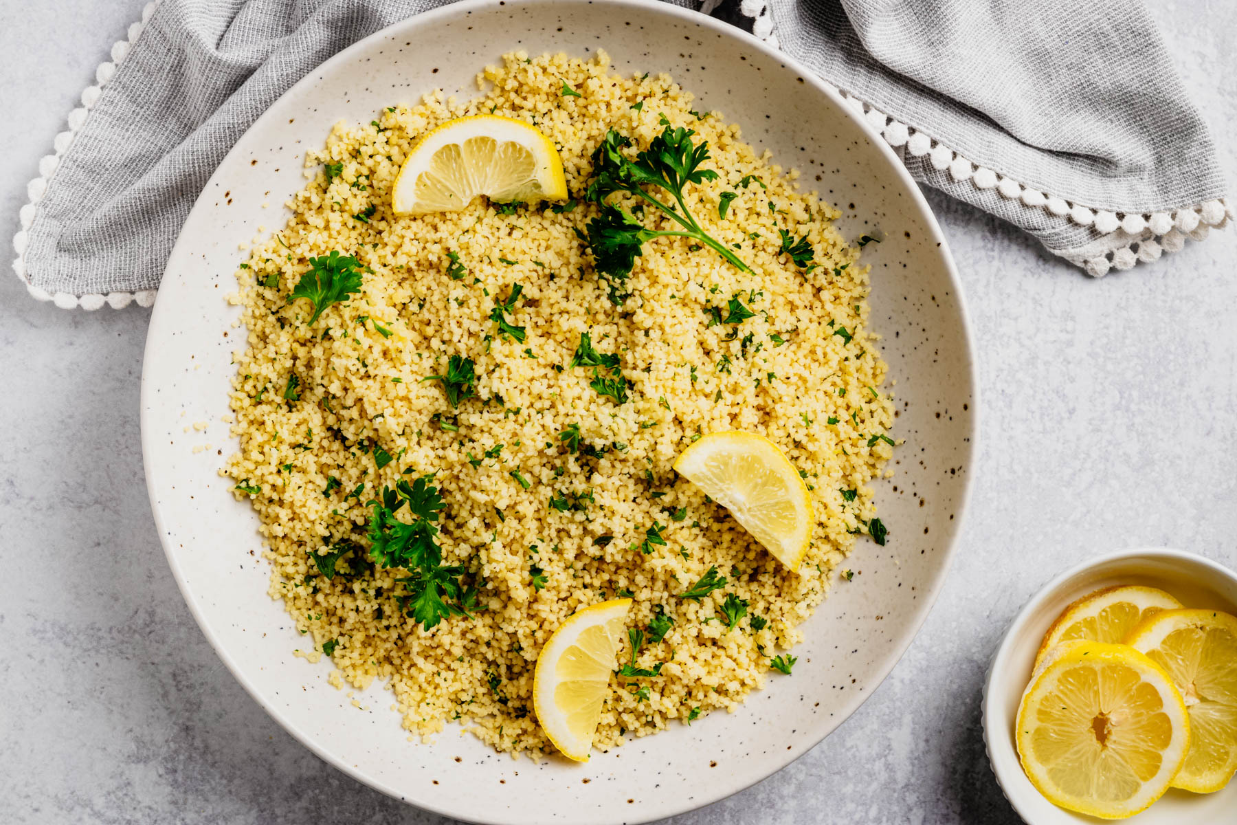 Overhead view of fluffy lemon couscous garnished with fresh parsley and lemon wedges in a large speckled serving bowl with sliced lemons on the side.