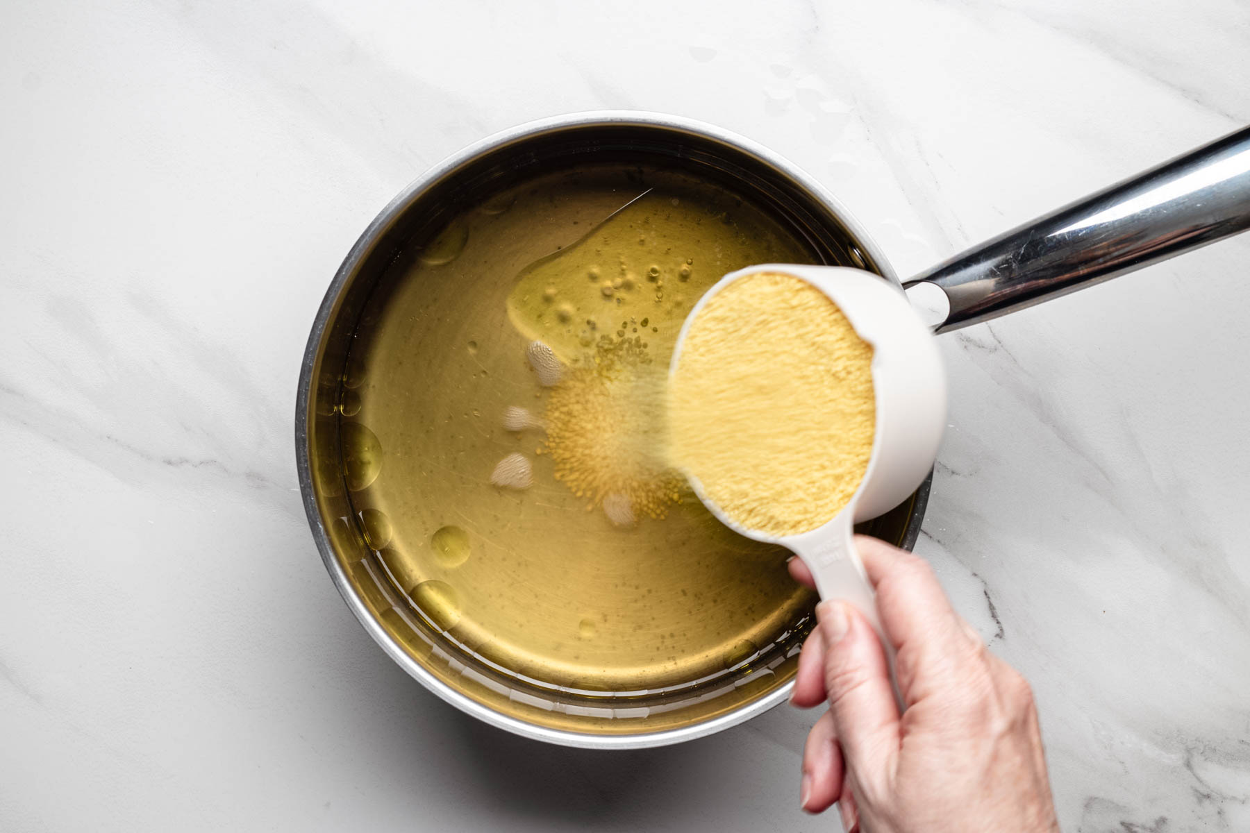 Fine couscous being poured from a measuring cup into hot broth in a saucepan.