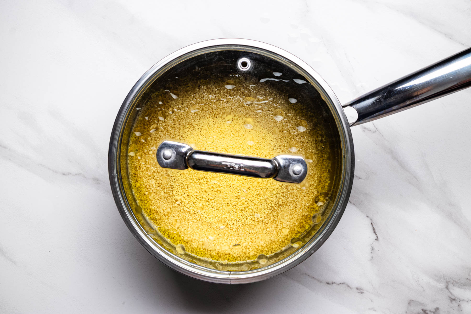Overhead view of couscous steaming in a covered saucepan after being added to hot broth and olive oil.