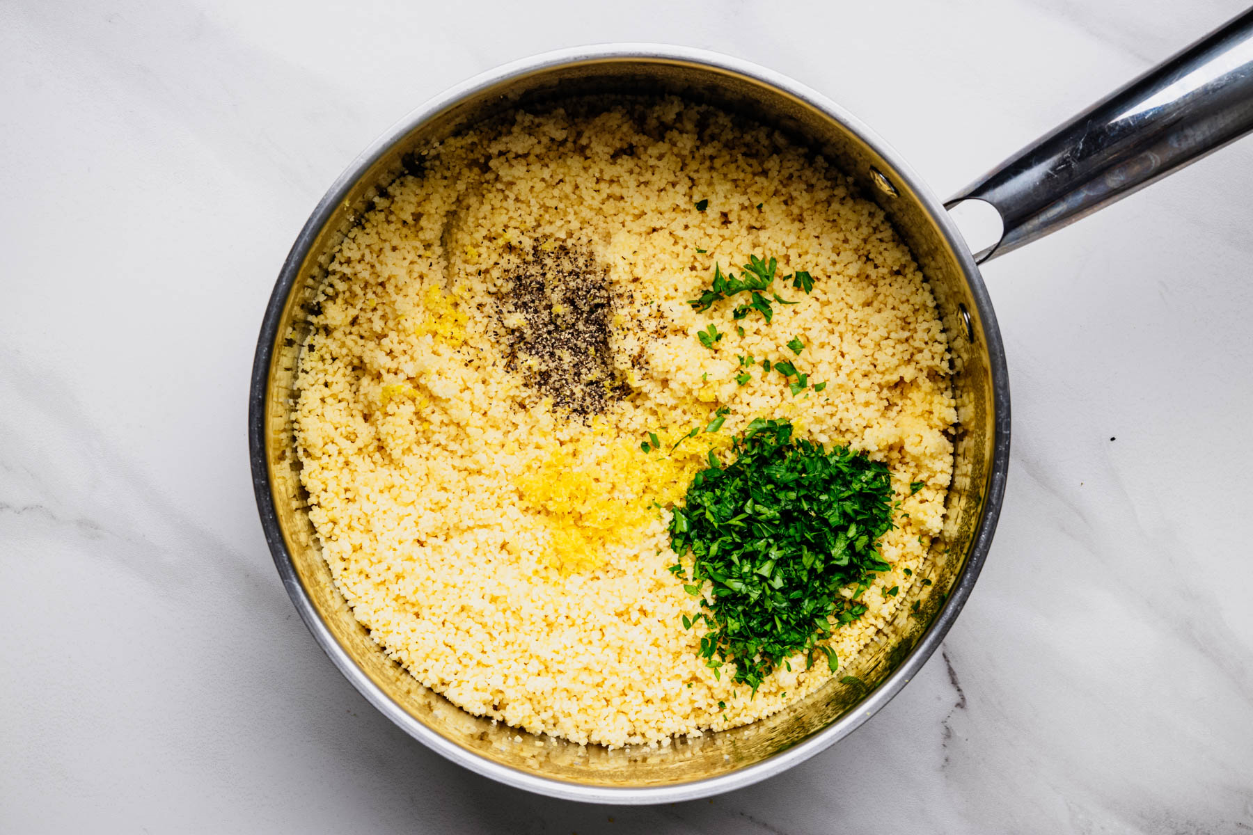 Fluffy cooked couscous in a saucepan topped with fresh parsley, lemon zest, and black pepper before mixing.
