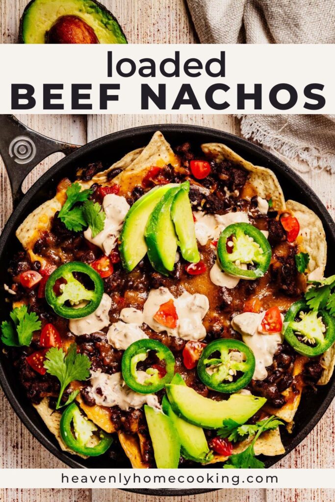 Skillet loaded beef nachos layered with seasoned beef, black beans, melted cheese, avocado slices, jalapeños, tomatoes, and cilantro. Vertical Pinterest pin for cheesy homemade beef nachos from Heavenly Home Cooking.