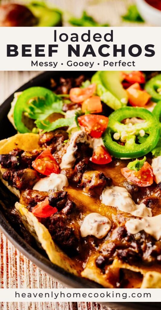 Loaded beef nachos topped with melted cheese, black beans, jalapeño slices, avocado, tomatoes, cilantro, and creamy sauce in a skillet. Vertical Pinterest pin for homemade loaded beef nachos from Heavenly Home Cooking.