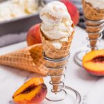 Homemade peach ice cream in a waffle cone with fresh peach slices and creamy frozen dessert in the background.