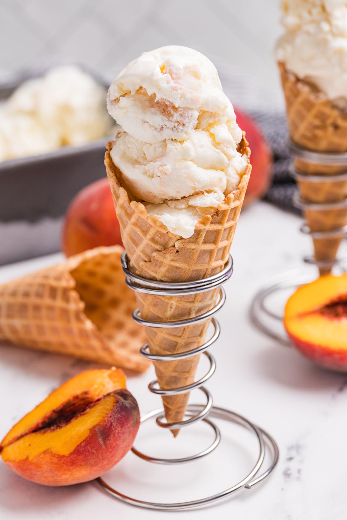 Close-up of creamy peach ice cream scoops in a waffle cone with fresh peaches and homemade ice cream nearby.