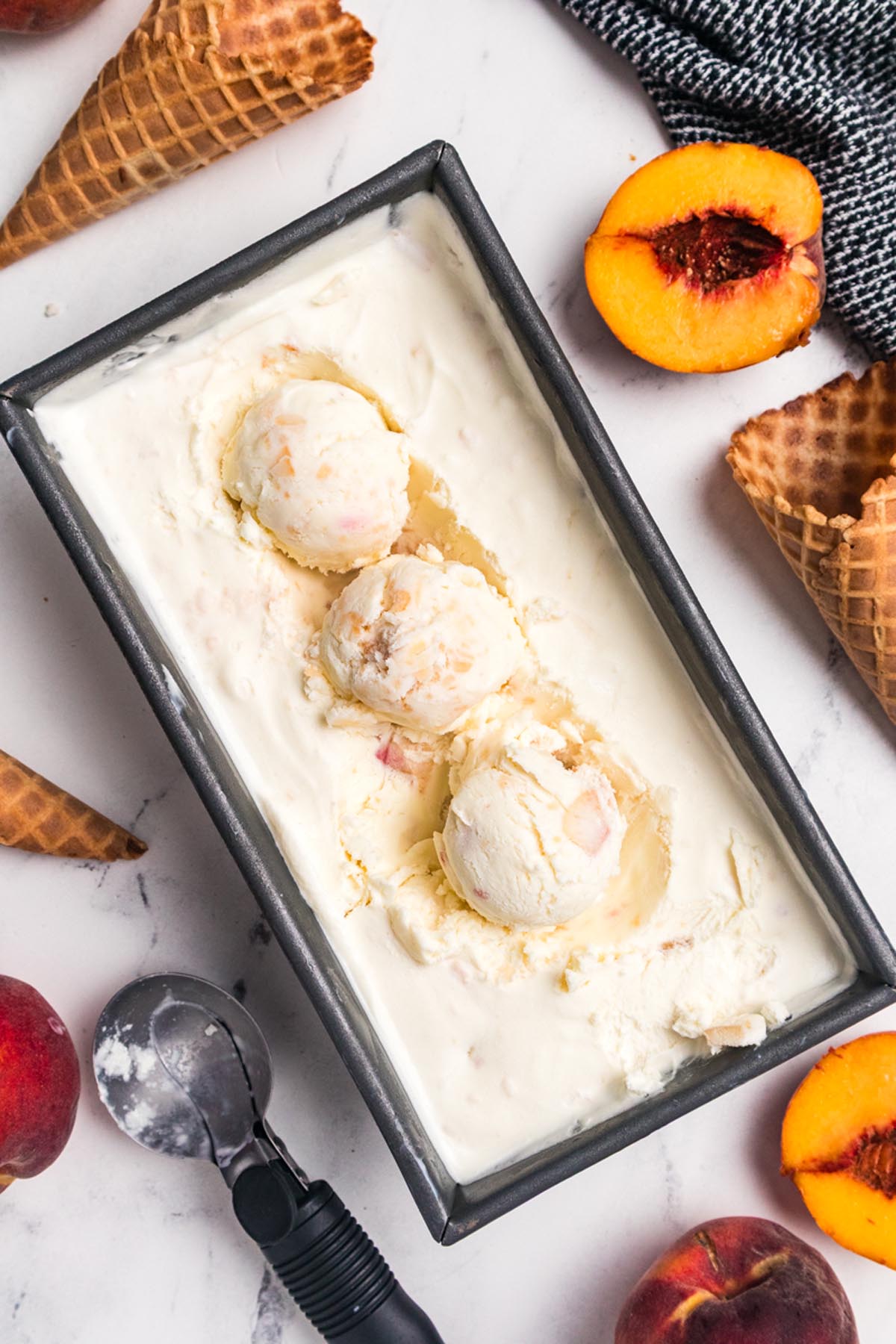 Homemade no churn peach ice cream scooped in a loaf pan with fresh peaches, waffle cones, and an ice cream scoop on a marble surface.