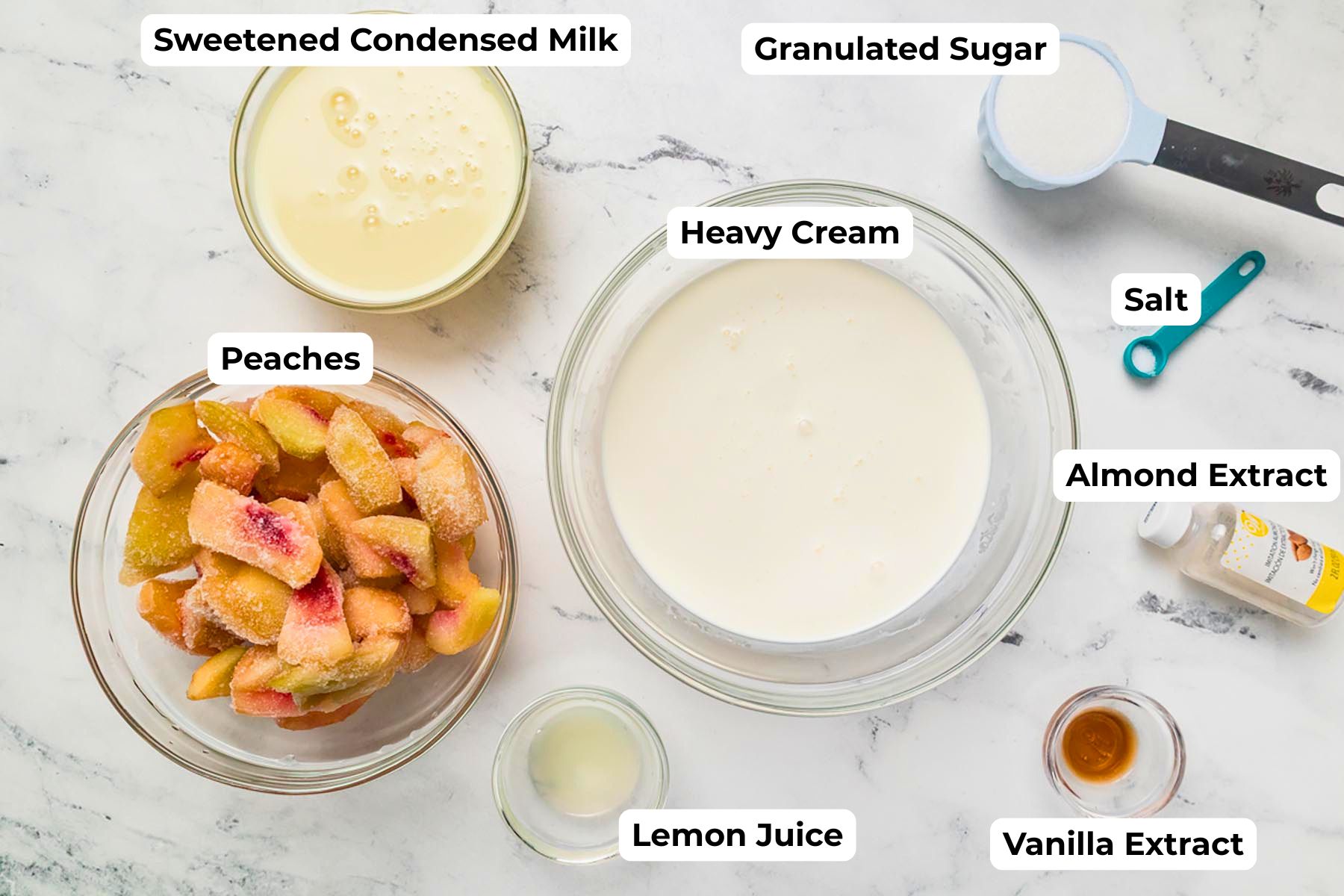Ingredients for homemade peach ice cream arranged on a marble surface including fresh peaches, heavy cream, sweetened condensed milk, sugar, vanilla, almond extract, lemon juice, and salt.