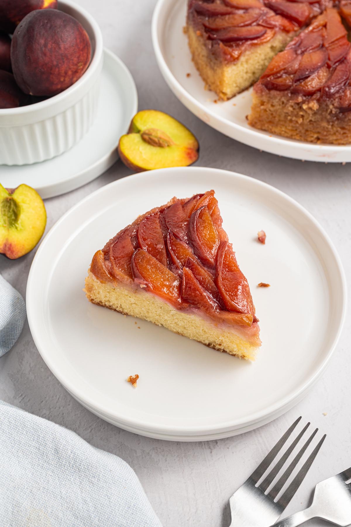 Slice of peach upside down cake served on a white plate with fresh peaches and the remaining cake in the background.