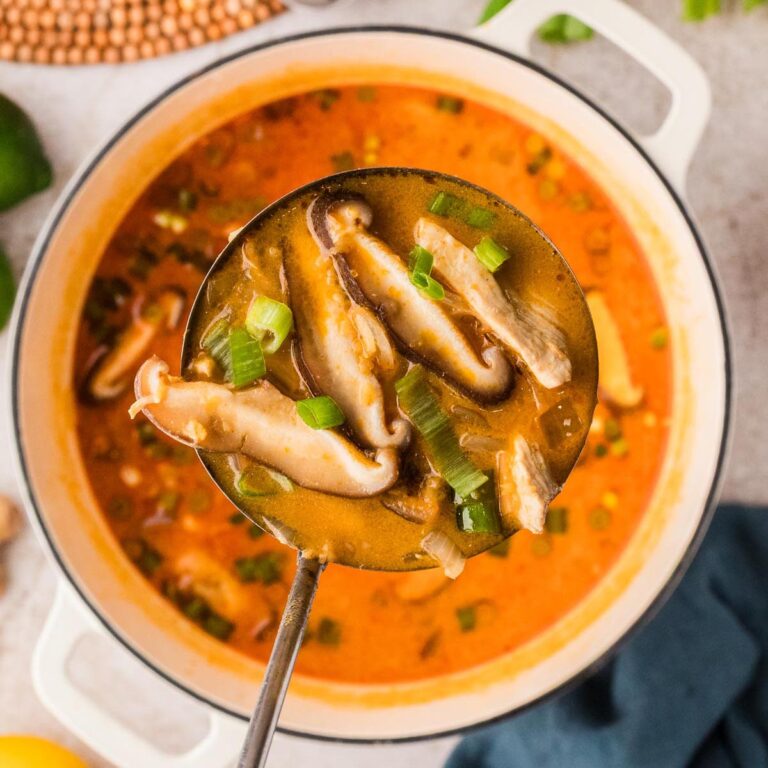 Thai chicken soup with red curry and coconut milk in a white Dutch oven, topped with green onions and mushrooms