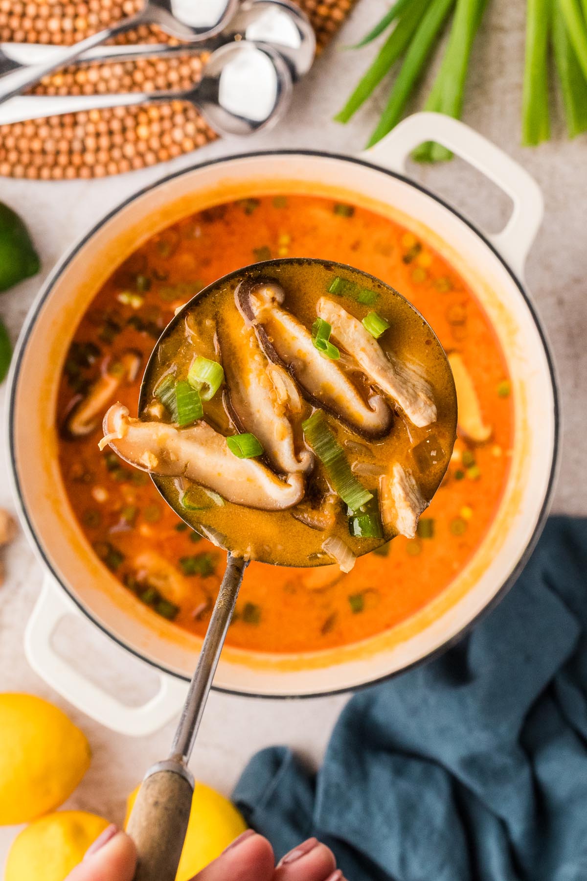 Ladle of Thai chicken curry soup with shiitake mushrooms and green onions over a pot of soup.
