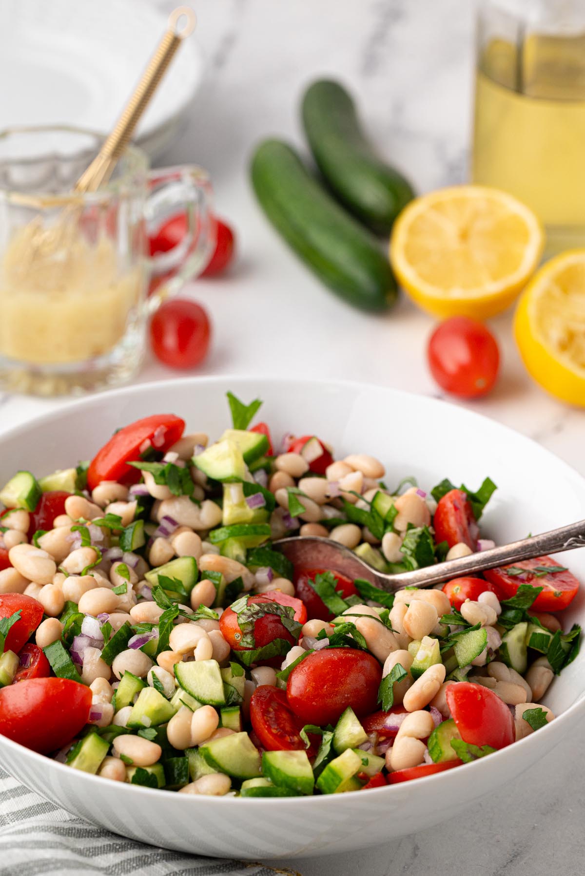 White bean salad with cherry tomatoes, cucumber, red onion, and fresh herbs in a white serving bowl with lemon vinaigrette ingredients in the background.