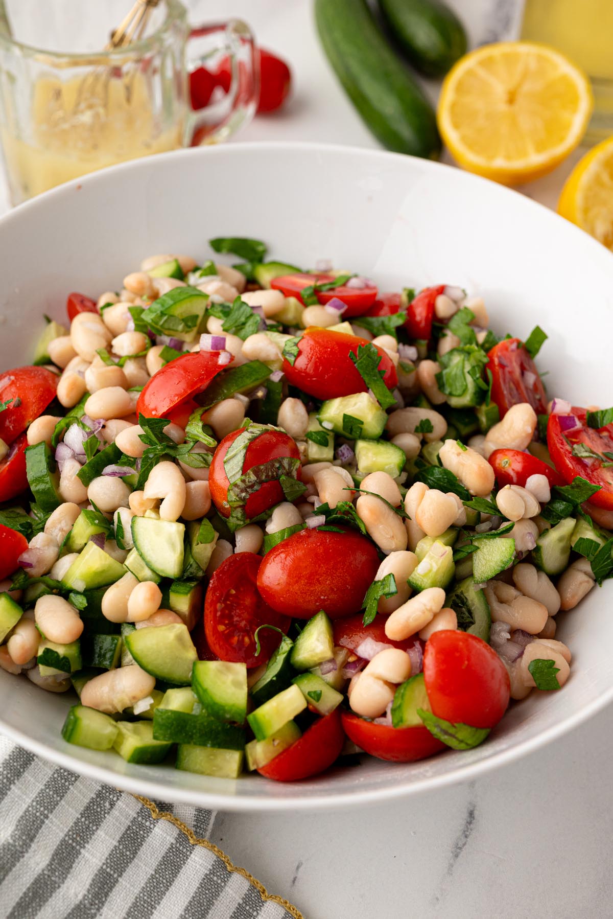 White bean salad with cherry tomatoes, cucumber, red onion, and fresh parsley in a white bowl with lemon vinaigrette ingredients in the background.