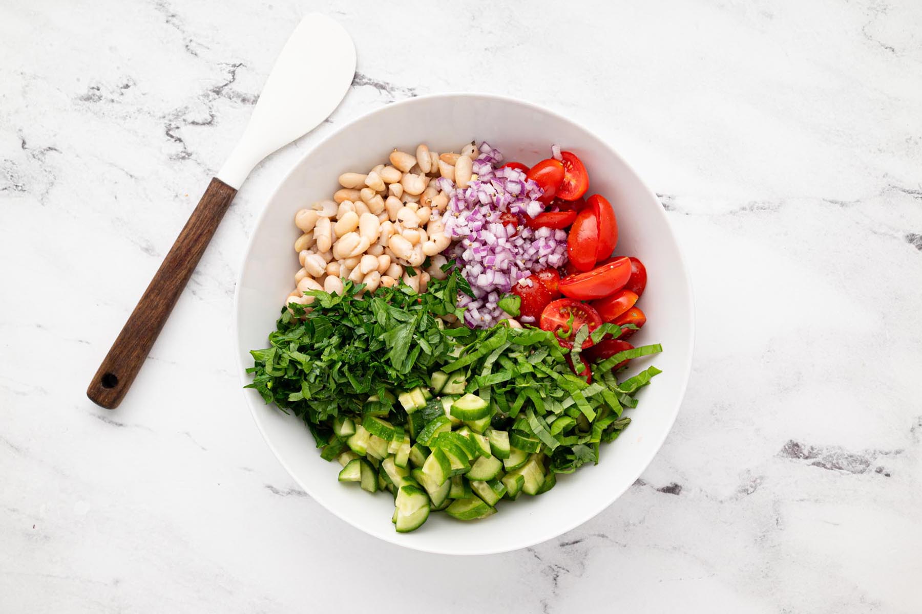 White beans, chopped cucumber, cherry tomatoes, red onion, parsley, and basil combined in a large white mixing bowl before adding dressing.