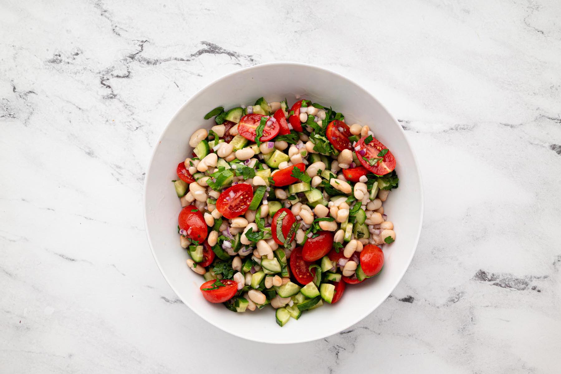 White bean salad with cherry tomatoes, cucumber, red onion, parsley, and basil served in a white bowl on a marble surface.