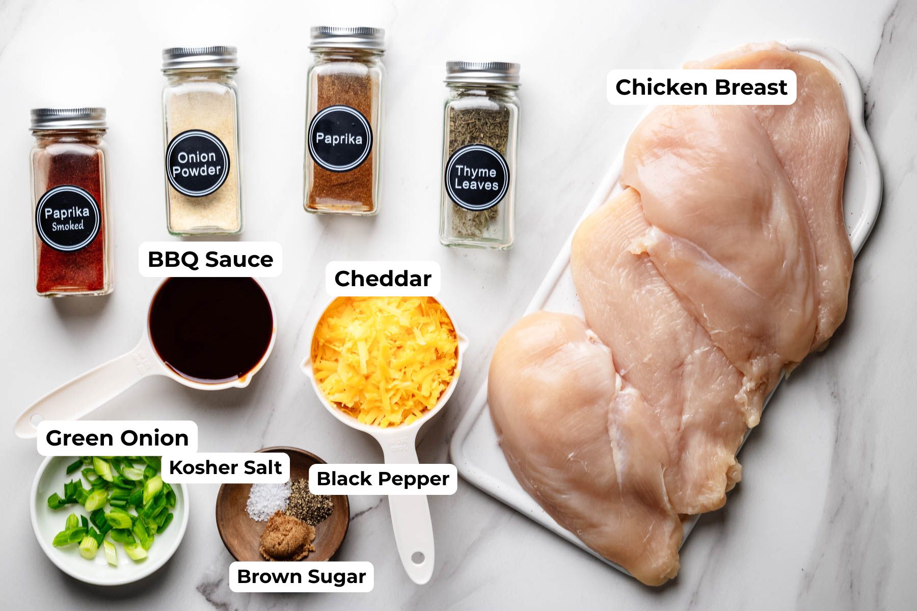 Ingredients for baked BBQ chicken breast arranged on a countertop, including chicken breasts, barbecue sauce, cheddar cheese, green onions, smoked paprika, paprika, onion powder, thyme, kosher salt, black pepper, and brown sugar.
