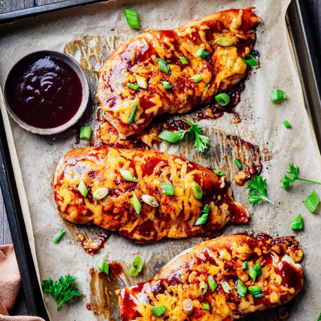 Baked BBQ chicken breasts topped with melted cheddar cheese and sliced green onions on a parchment-lined sheet pan, served with a bowl of barbecue sauce.