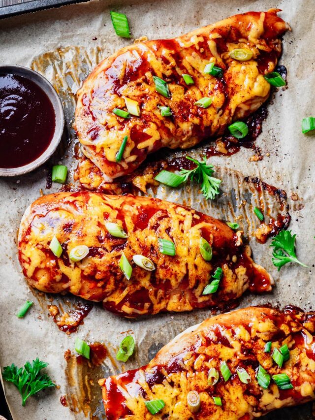 Baked BBQ chicken breasts topped with melted cheddar cheese and sliced green onions on a parchment-lined sheet pan, served with a bowl of barbecue sauce.