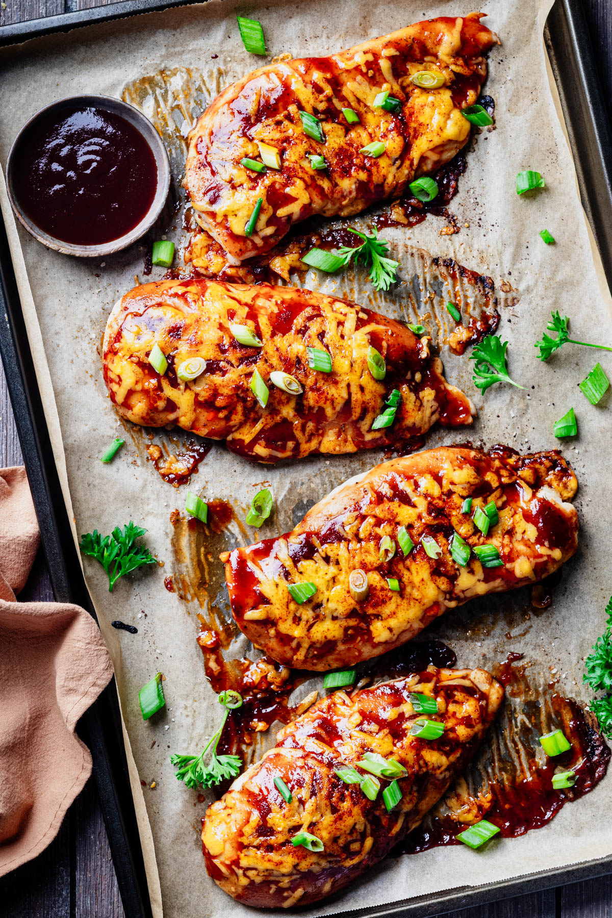 Four baked BBQ chicken breast cutlets topped with cheddar cheese and sliced scallions on a parchment-lined sheet pan with a bowl of barbecue sauce.