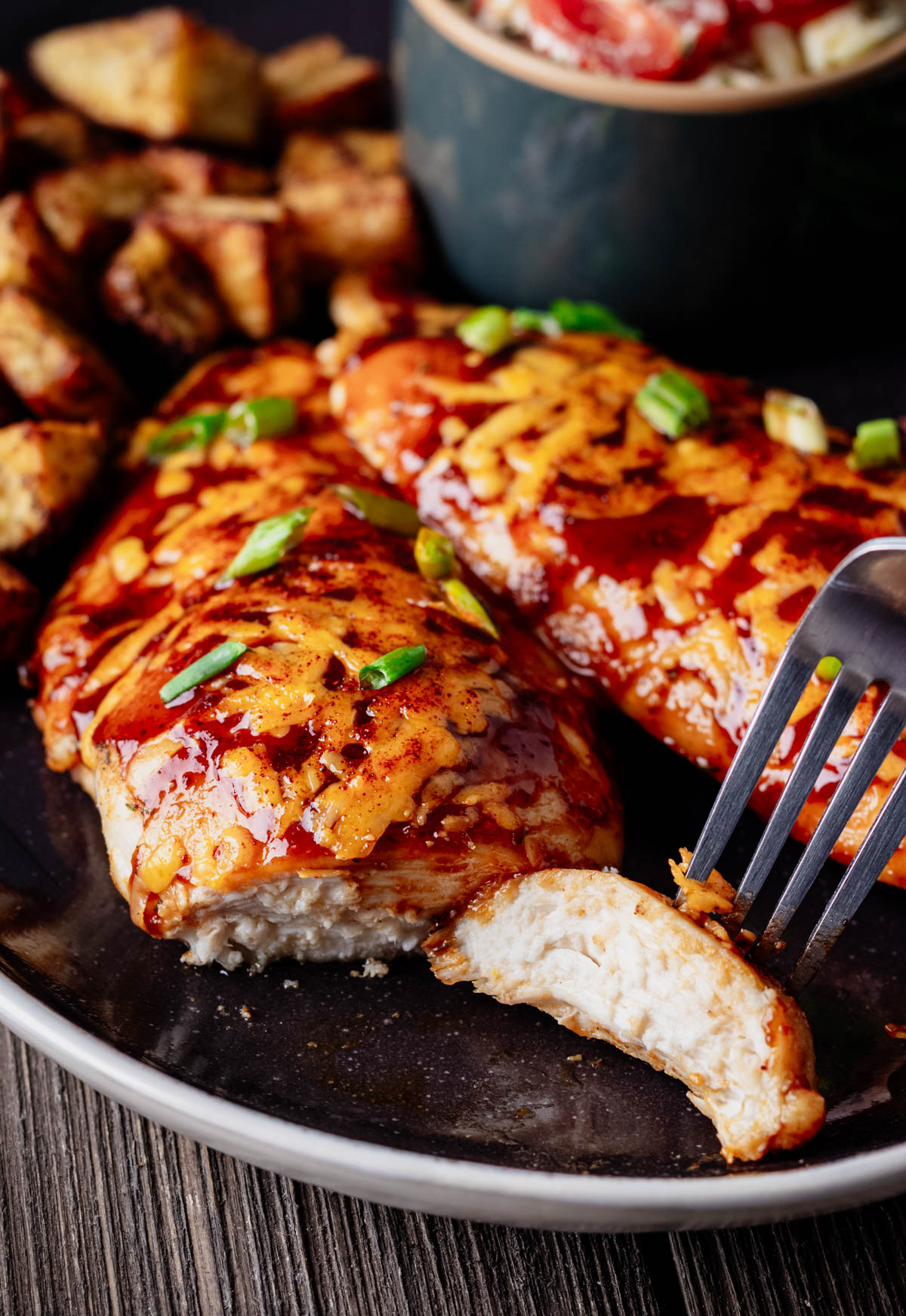 Close-up of baked BBQ chicken breast cutlets topped with melted cheddar cheese, sliced to show the juicy interior, and garnished with green onions. Roasted potatoes and potato salad are blurred in the background.