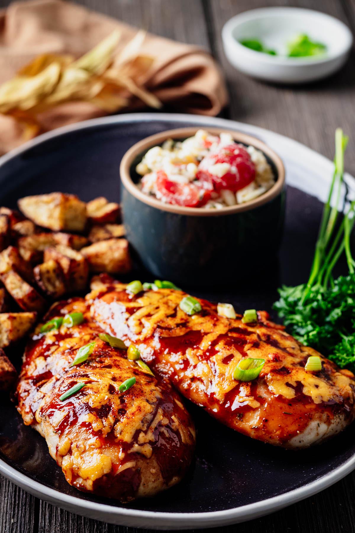 Baked BBQ chicken breast served with roasted potatoes and pasta salad on a dark dinner plate.