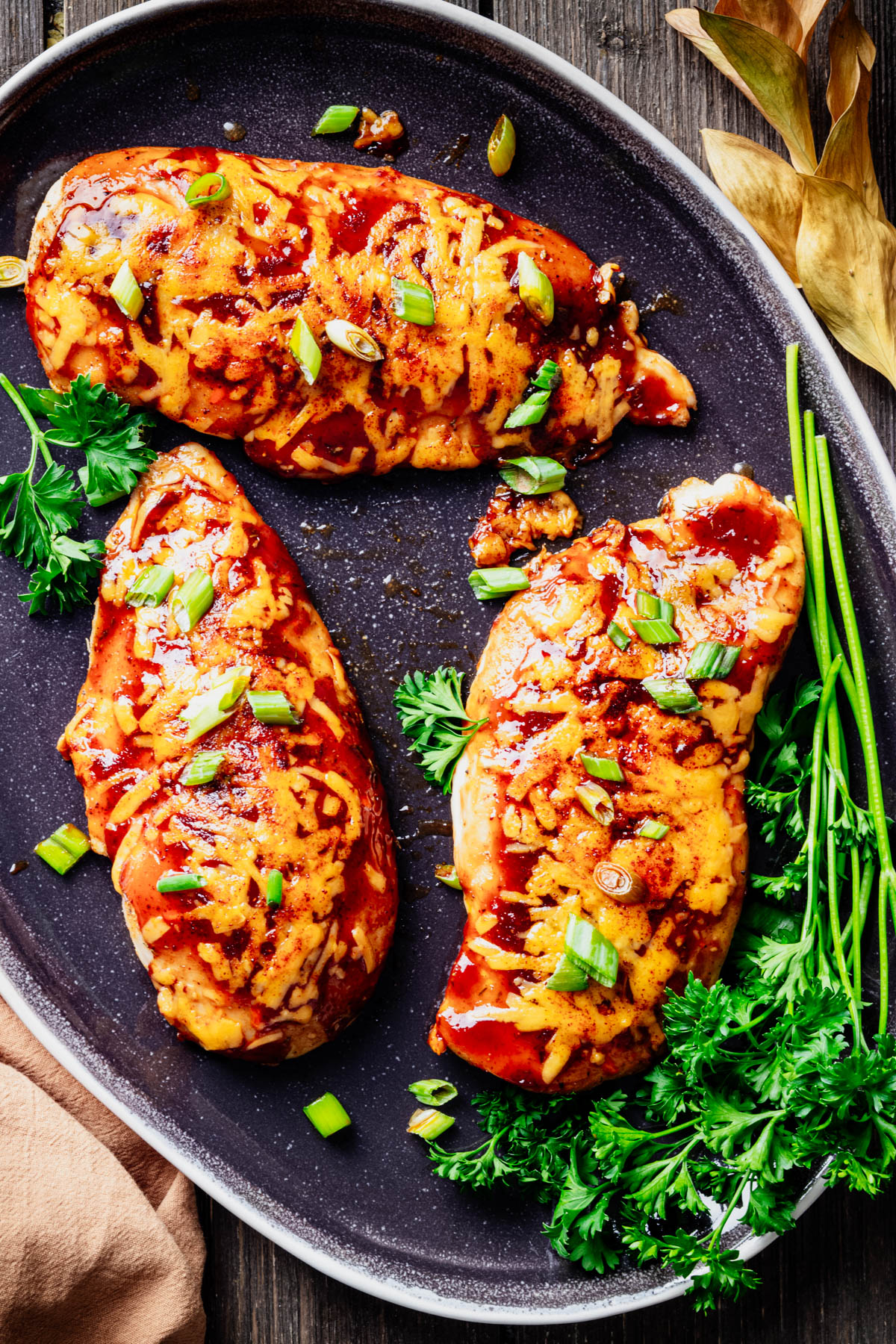 Three baked BBQ chicken breasts topped with melted cheddar cheese and sliced green onions, arranged on a dark serving platter and garnished with fresh parsley.