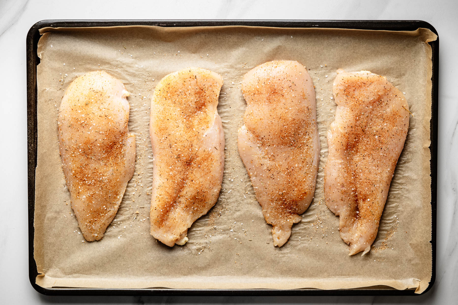 Raw chicken breast cutlets arranged on a parchment-lined baking sheet and seasoned with a blend of spices.