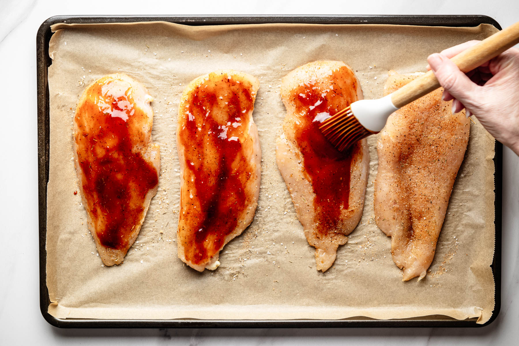Barbecue sauce being brushed over seasoned chicken breast cutlets on a parchment-lined baking sheet.