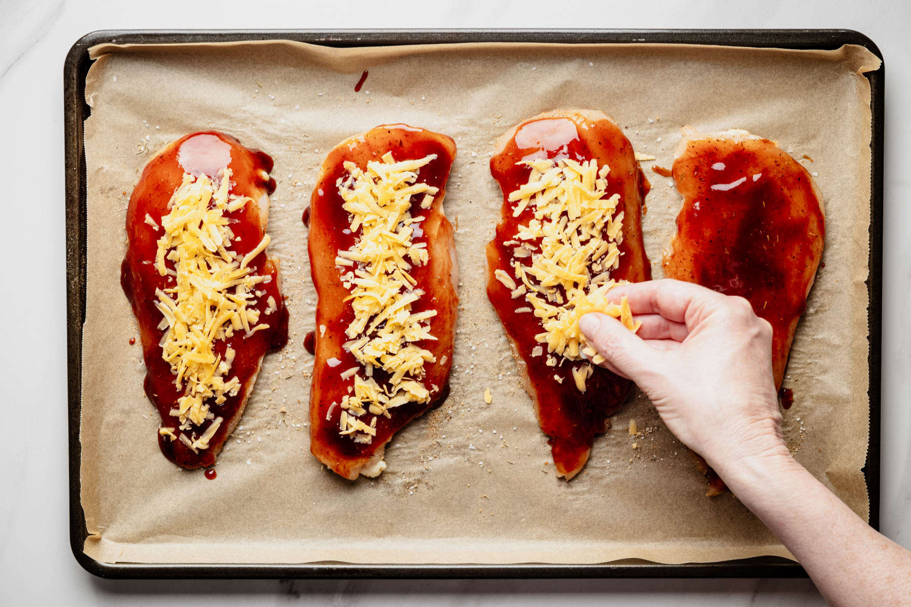Shredded cheddar cheese being sprinkled over barbecue sauce-coated chicken breast cutlets before baking.