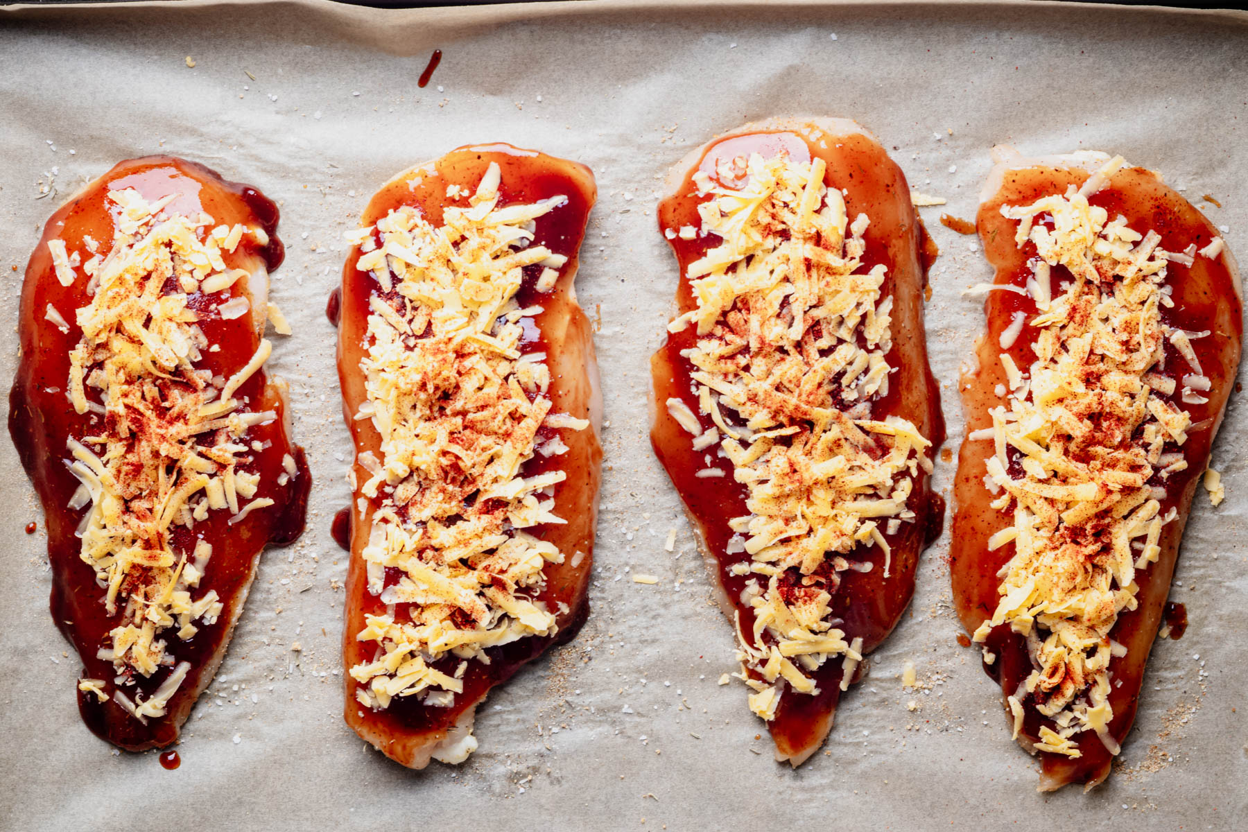 BBQ chicken breast cutlets topped with cheddar cheese and paprika, ready to bake on a parchment-lined baking sheet.