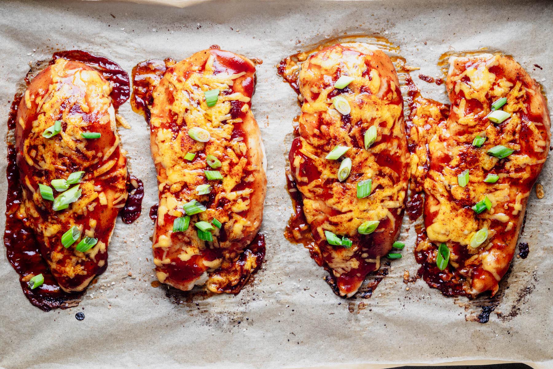 Baked BBQ chicken breast cutlets topped with melted cheddar cheese, barbecue sauce, and sliced green onions on a baking sheet.