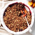 Blueberry peach crisp in a white baking dish with a spoon lifting a serving to reveal the juicy peach and blueberry filling beneath the oat crumble topping.
