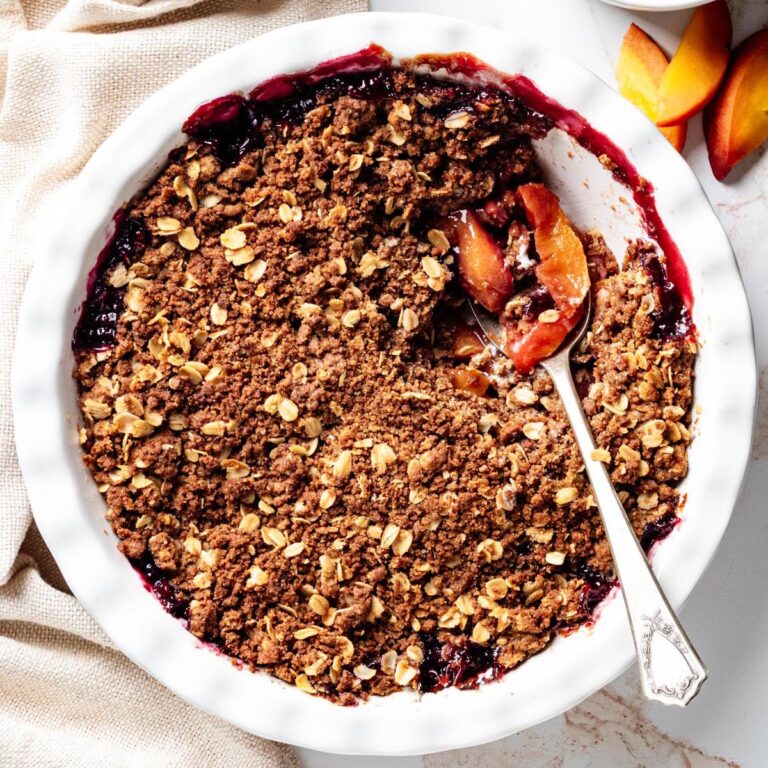 Blueberry peach crisp in a white baking dish with a spoon lifting a serving to reveal the juicy peach and blueberry filling beneath the oat crumble topping.