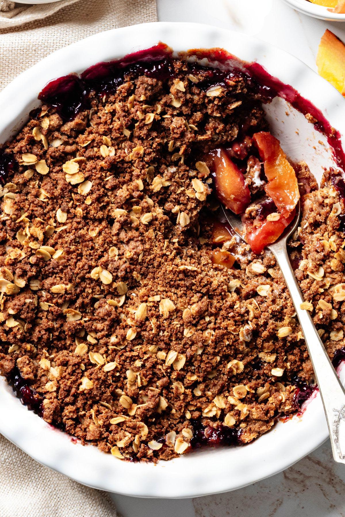 Overhead view of a blueberry peach crisp with a ginger oat topping in a white baking dish. A spoon scoops out a serving, revealing the juicy peach and blueberry filling beneath the crisp topping.