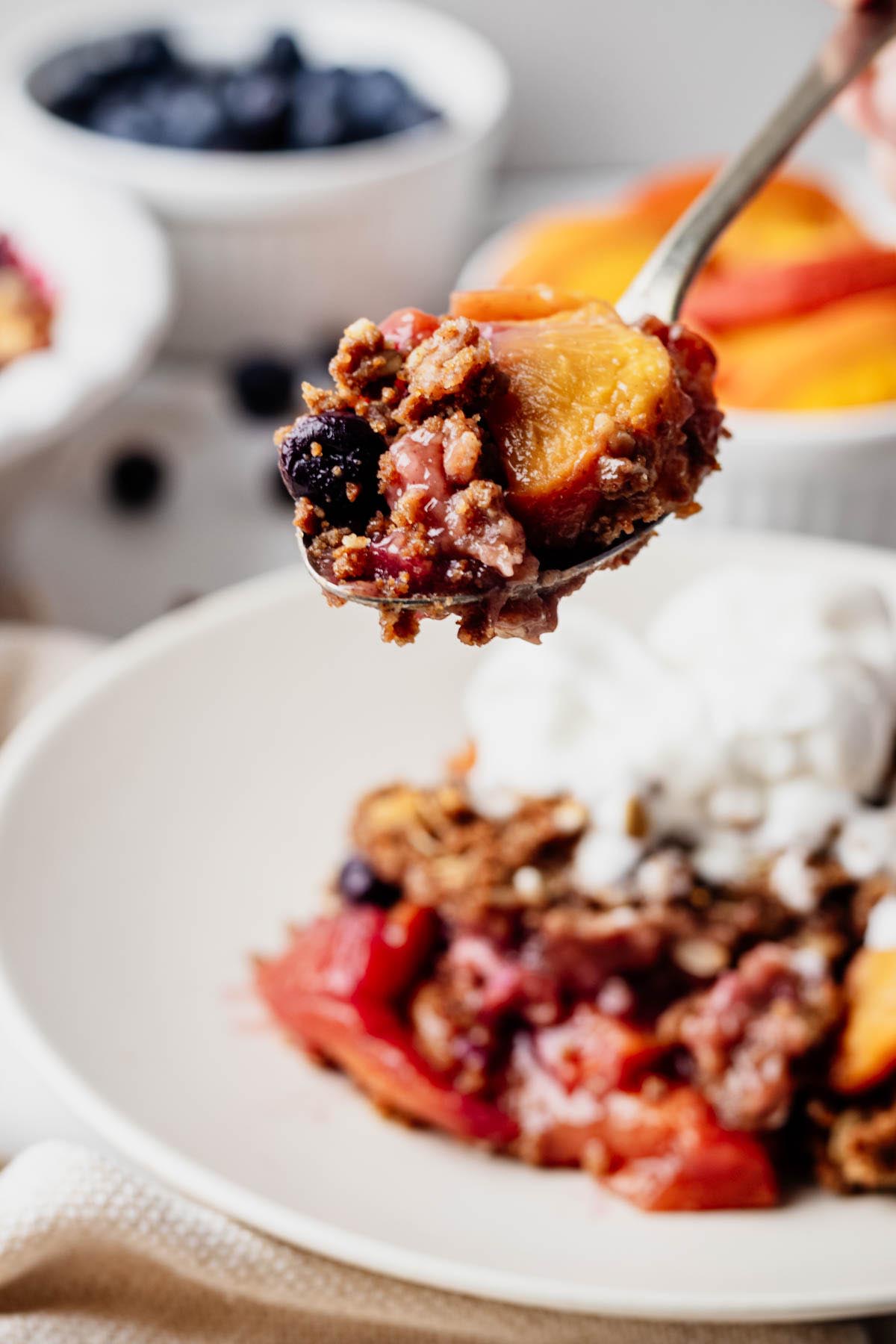 Spoonful of blueberry peach crisp held above a bowl of crisp topped with vanilla ice cream.