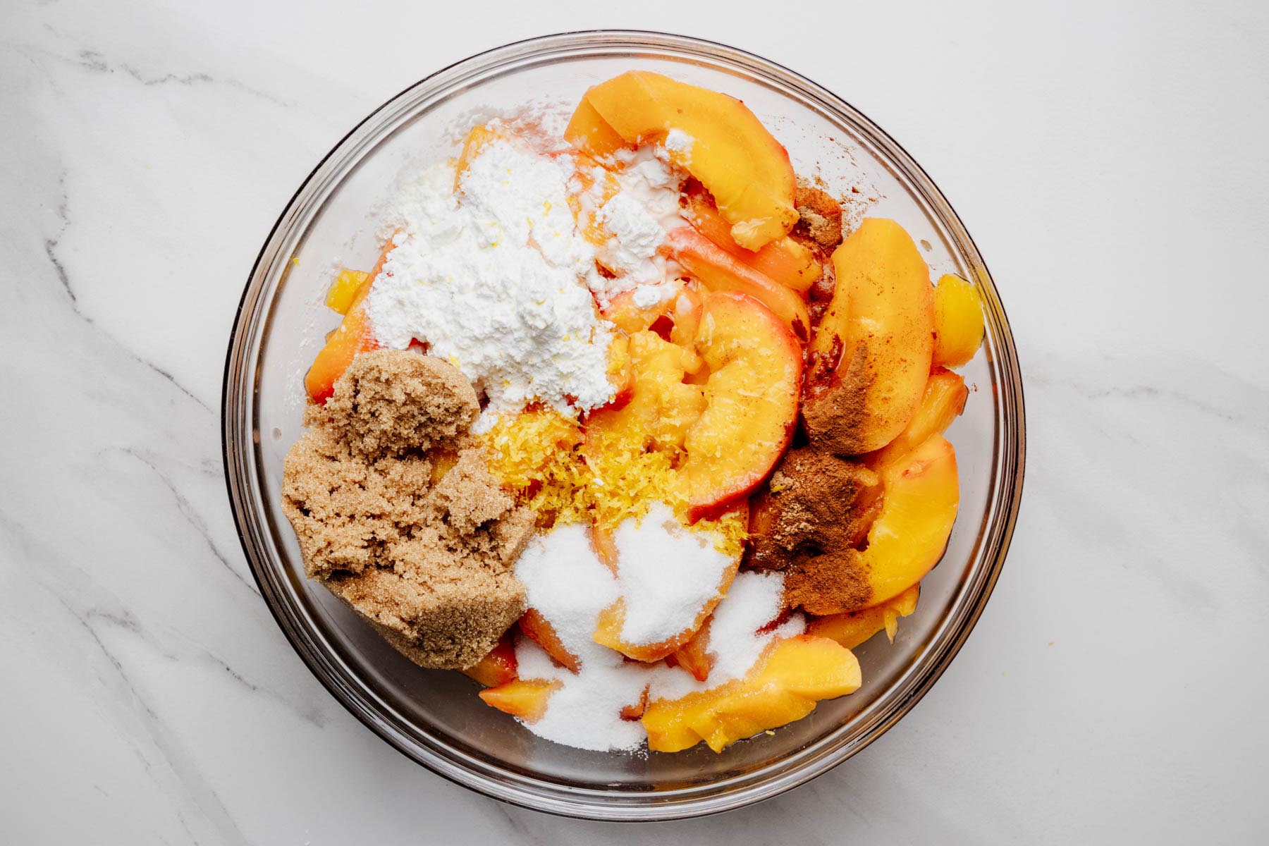 Sliced peaches in a glass bowl with brown sugar, granulated sugar, cornstarch, lemon zest, cinnamon, ginger, and other filling ingredients.