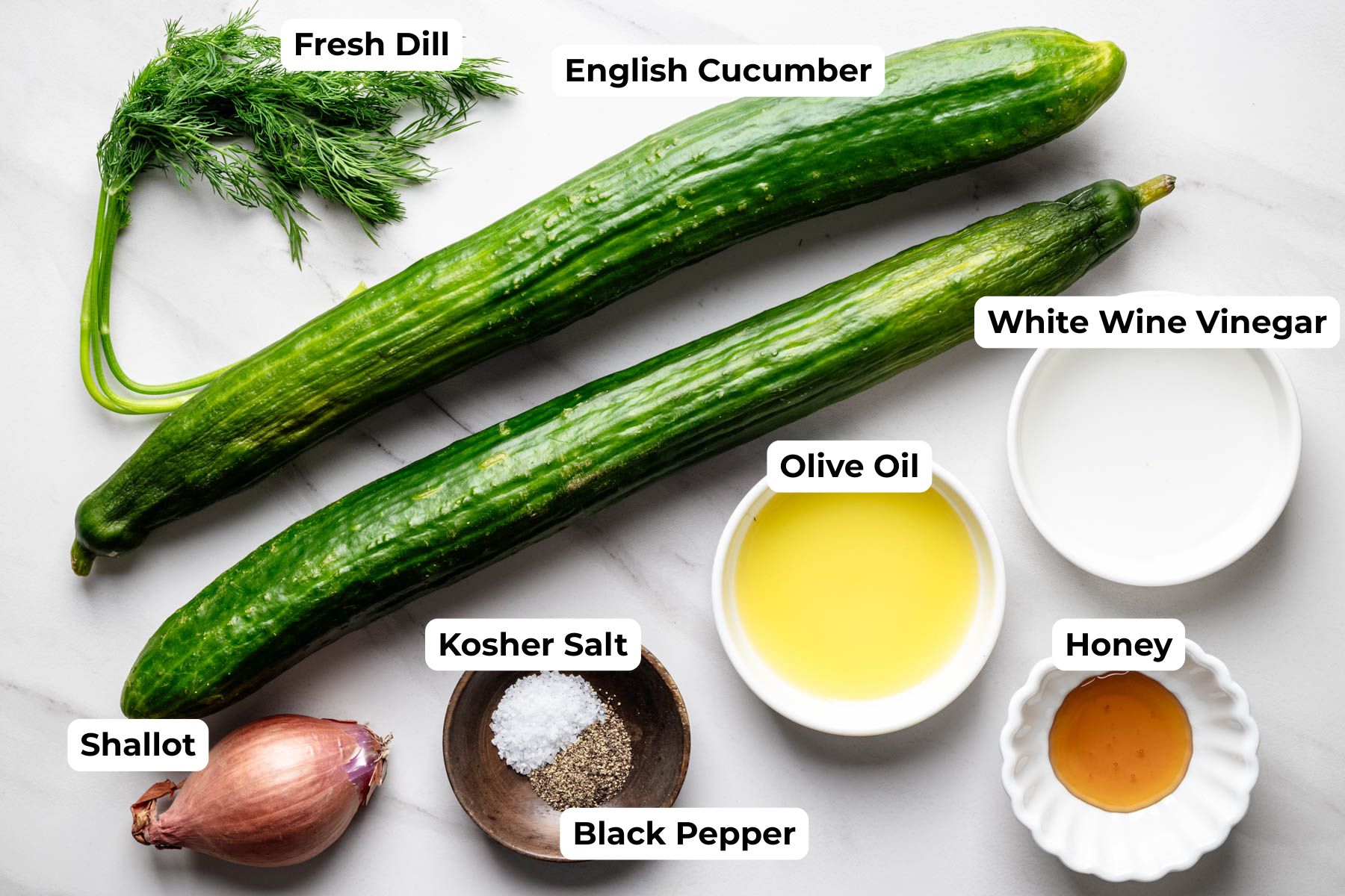 Labeled ingredients for cucumber dill salad, including English cucumbers, fresh dill, shallot, olive oil, white wine vinegar, honey, kosher salt, and black pepper arranged on a white marble surface.