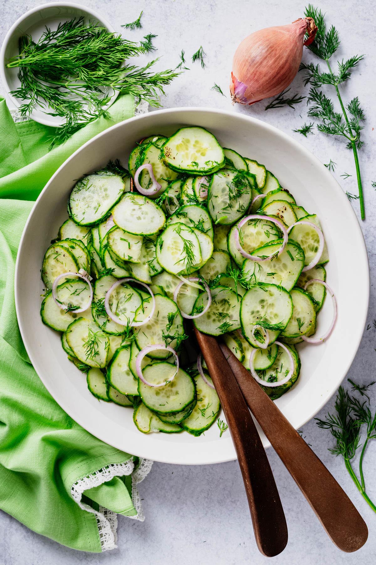 English cucumber dill salad with fresh dill and shallot tossed in a light white wine vinaigrette, served in a white bowl with wooden serving spoons.