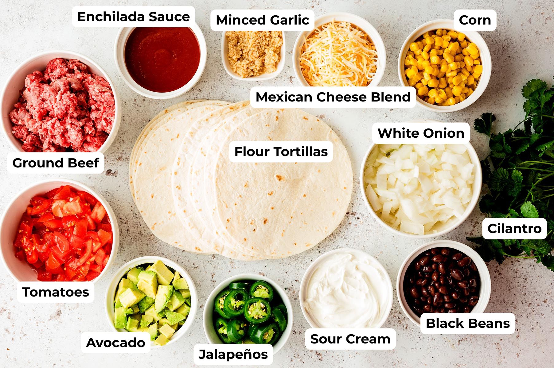 Ingredients for enchilada cups arranged on a light countertop, including flour tortillas, ground beef, enchilada sauce, Mexican cheese blend, corn, white onion, black beans, sour cream, tomatoes, avocado, jalape&ntilde;os, minced garlic, and fresh cilantro.