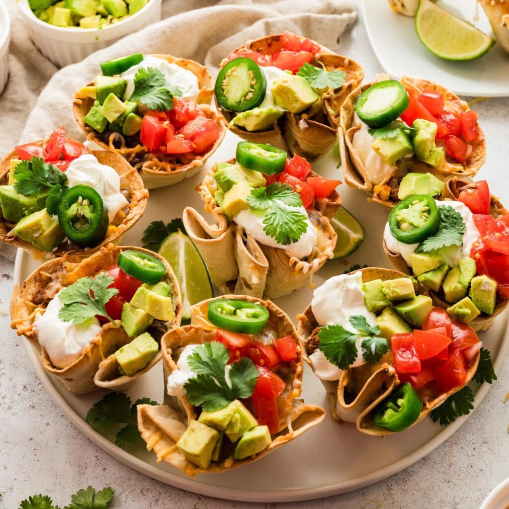 Platter of enchilada cups topped with diced avocado, tomatoes, jalape&ntilde;o slices, sour cream, and fresh cilantro, served with lime wedges.