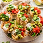 Platter of enchilada cups topped with diced avocado, tomatoes, jalapeño slices, sour cream, and fresh cilantro, served with lime wedges.