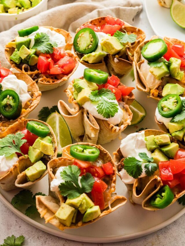 Platter of enchilada cups topped with diced avocado, tomatoes, jalape&ntilde;o slices, sour cream, and fresh cilantro, served with lime wedges.