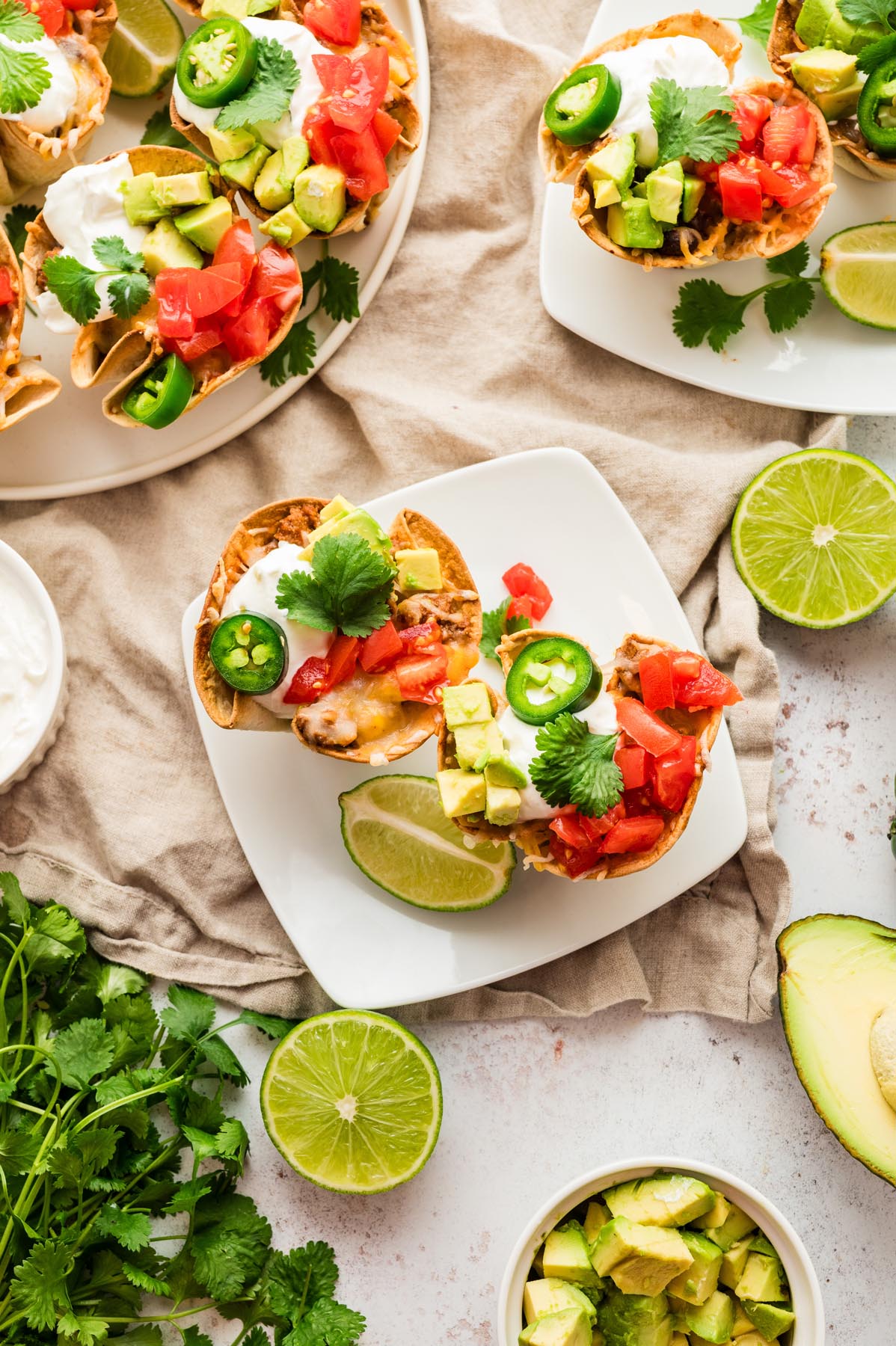 Two enchilada cups topped with avocado, tomatoes, jalape&ntilde;os, sour cream, and cilantro served on a white plate with lime wedges.