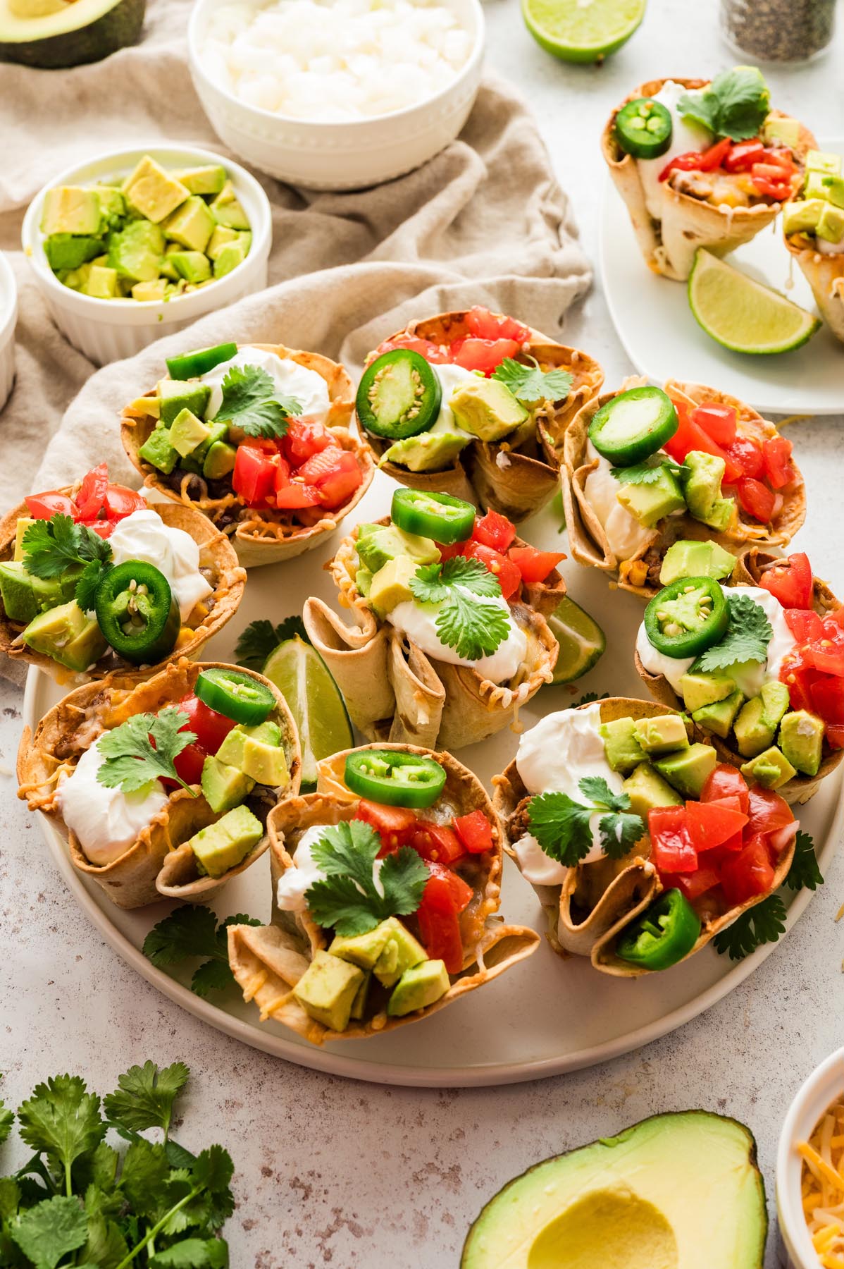 Plate of enchilada cups topped with avocado, tomatoes, jalape&ntilde;os, sour cream, and cilantro, served with lime wedges.