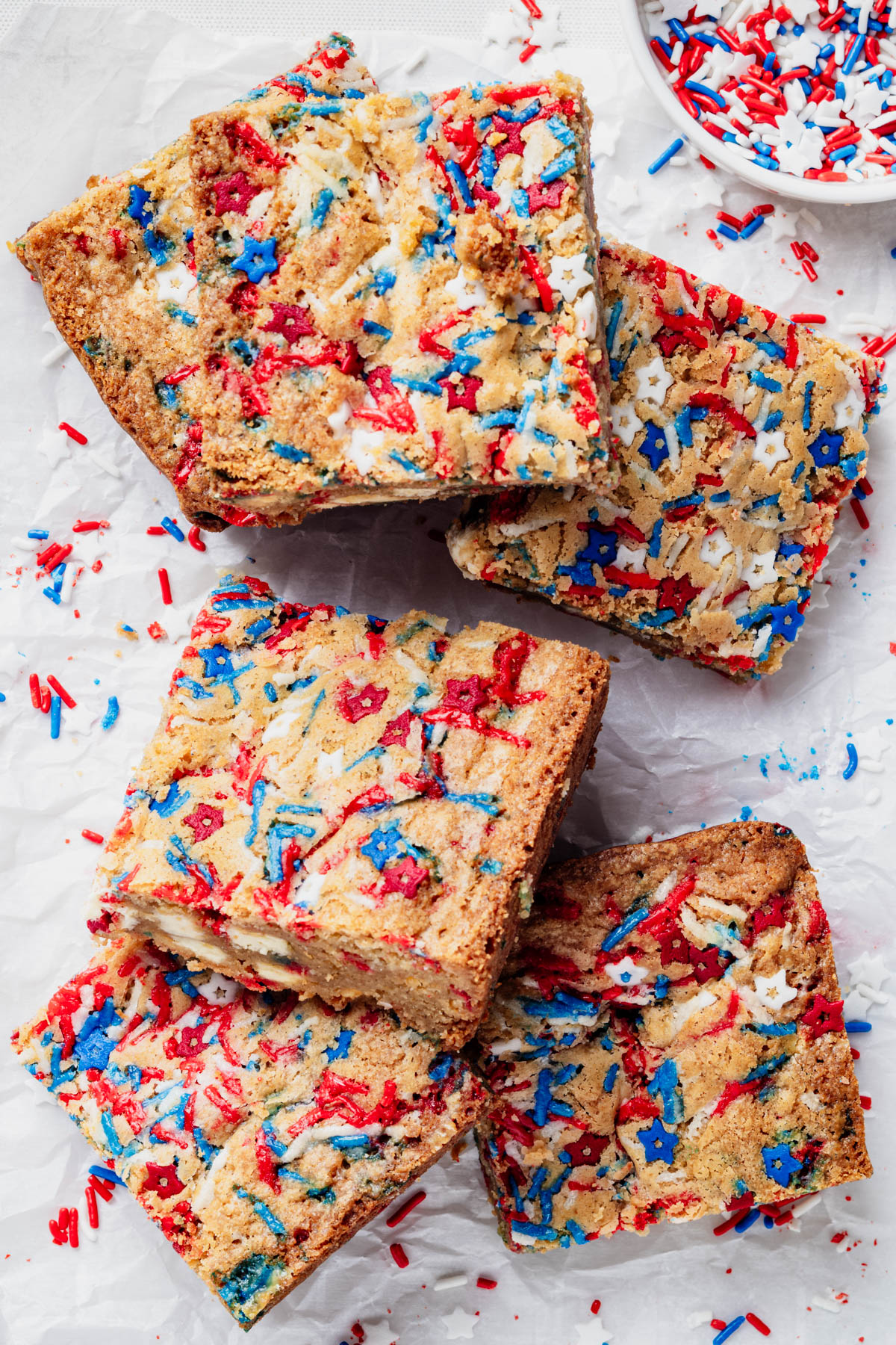 Overhead view of fireworks blondies with white chocolate chunks and patriotic red, white, and blue sprinkles arranged on parchment paper.