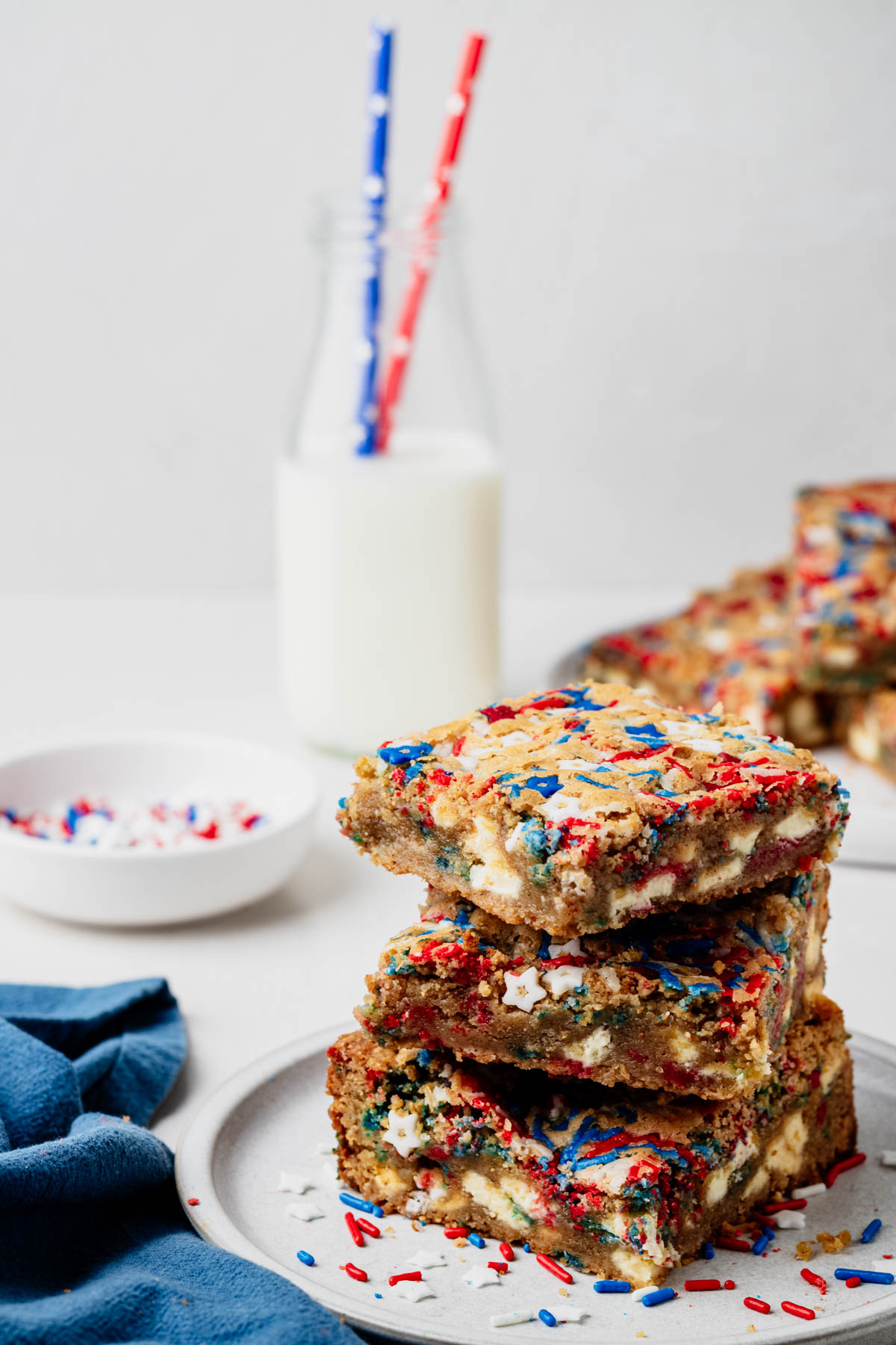Three Fireworks Blondies stacked on a gray plate, topped with red, white, and blue sprinkles and filled with white chocolate chunks, with milk and additional blondies in the background.