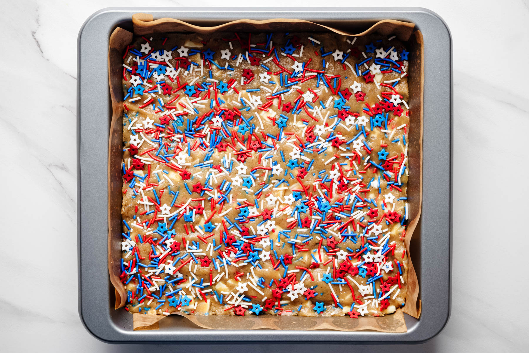 Blondie batter spread into a parchment-lined baking pan and topped with red, white and blue sprinkles.