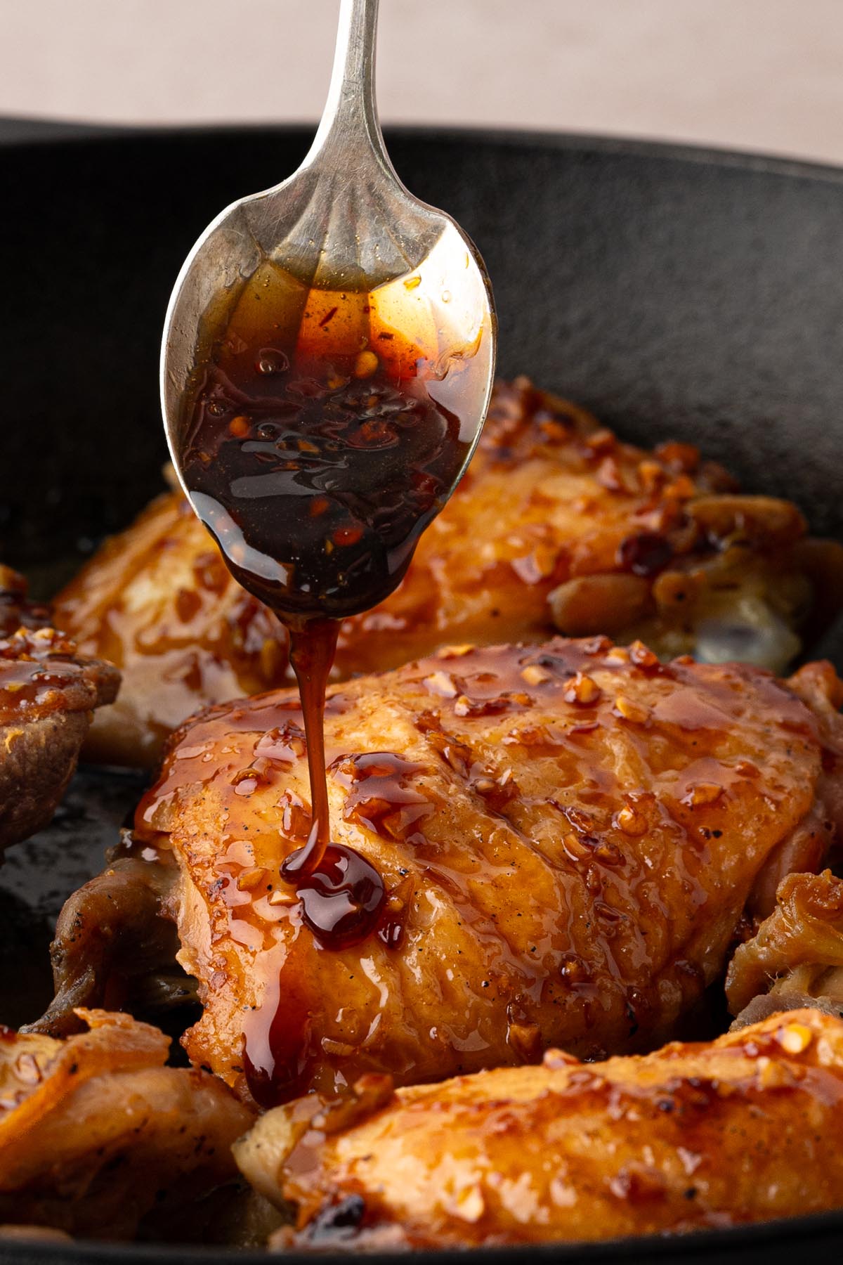 A spoon drizzles sticky honey garlic sauce over chicken thighs in a skillet during the final glazing step.