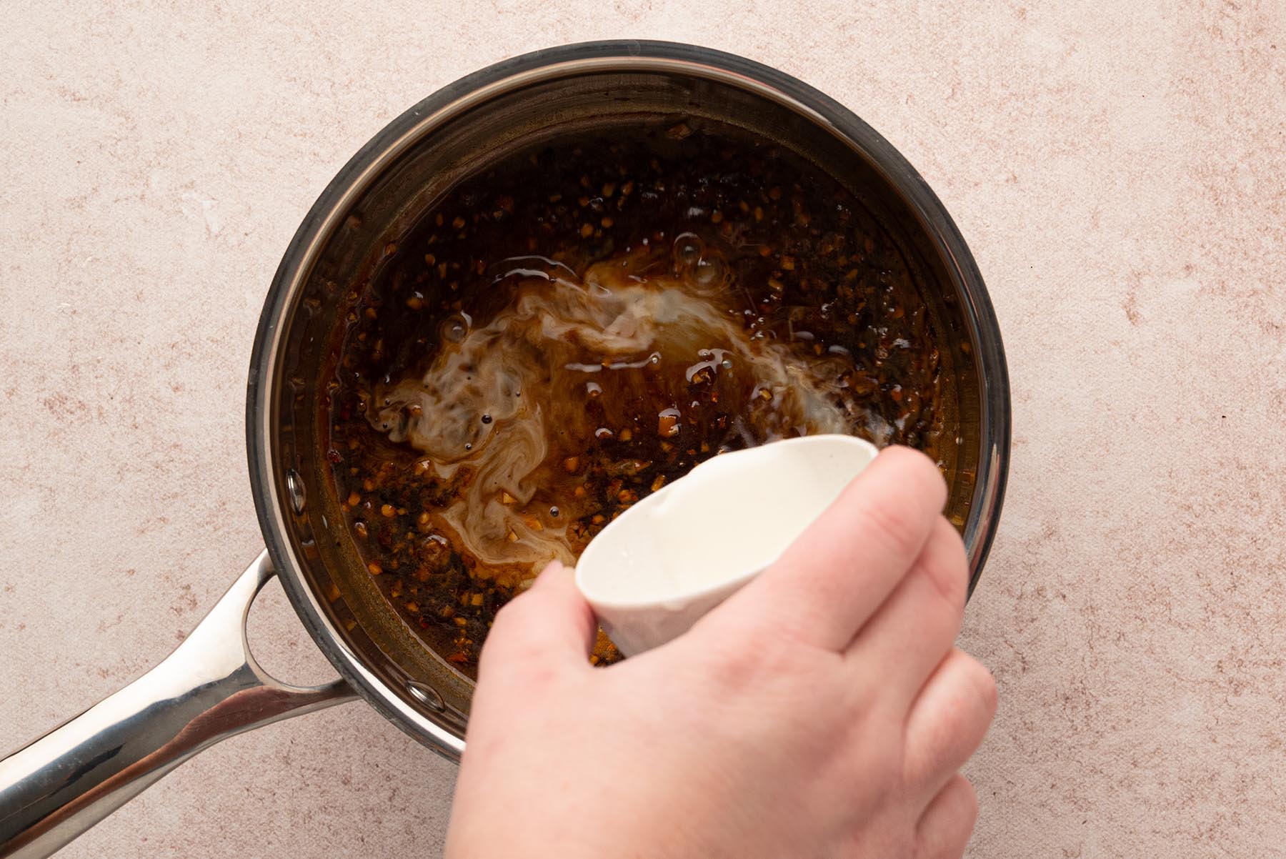 An overhead view of a cornstarch slurry being poured into the honey garlic sauce in a saucepan.