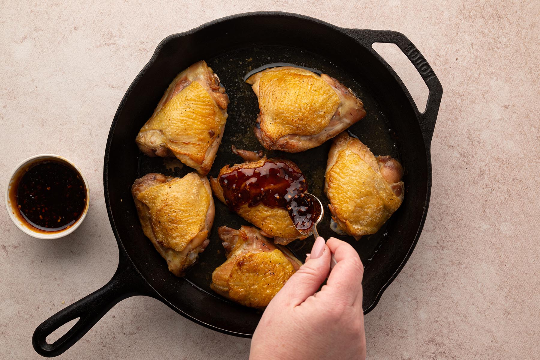 An overhead view of honey garlic sauce being spooned over baked chicken thighs in a cast iron skillet.