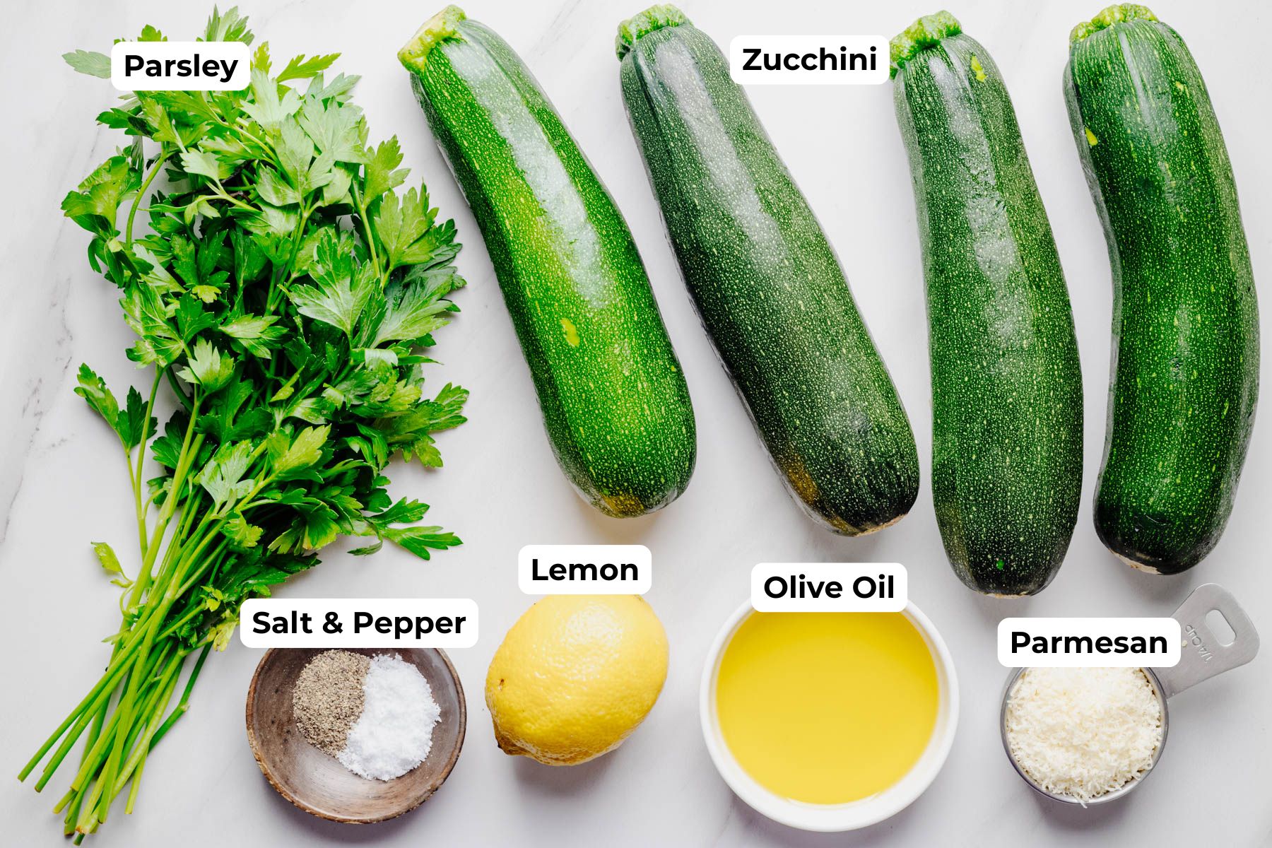 Fresh parsley, whole zucchini, olive oil, a lemon, grated Parmesan cheese, and salt and pepper arranged on a countertop with labels identifying each ingredient for Parmesan grilled zucchini.