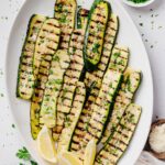 Overhead view of grilled zucchini arranged on a white serving platter and topped with grated Parmesan cheese and chopped fresh parsley. Lemon wedges are tucked alongside the zucchini, with crisp grill marks visible on every slice.